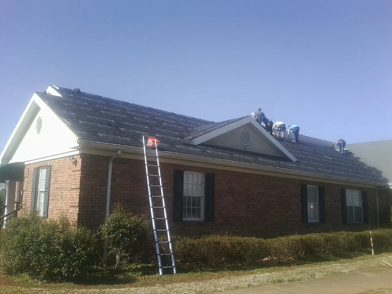 Crew tearing shingles off a brick ranch roof with a ladder set — Metal Roof Repair work by our crew in Chilo