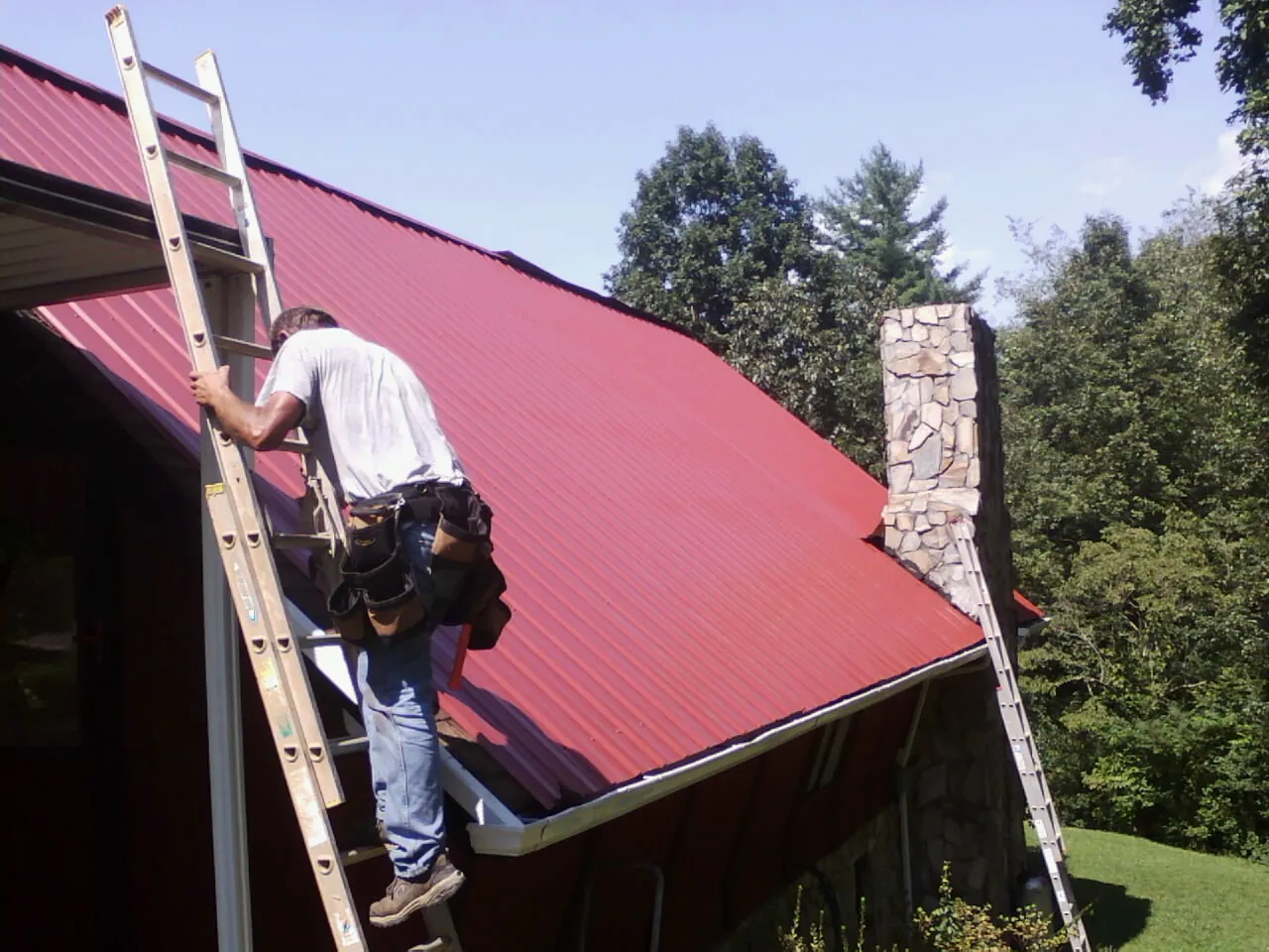 Roofer on a ladder beside a red metal roof and stone chimney — Soffit & Fascia Repair by Roofing Brothers in Chilo